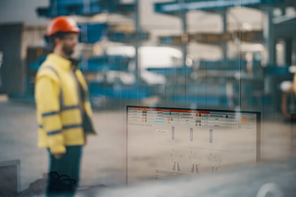 Worker in safety gear monitoring drilling system data displayed on a screen in a workshop. Worker in safety gear monitoring drilling system data displayed on a screen in a workshop.