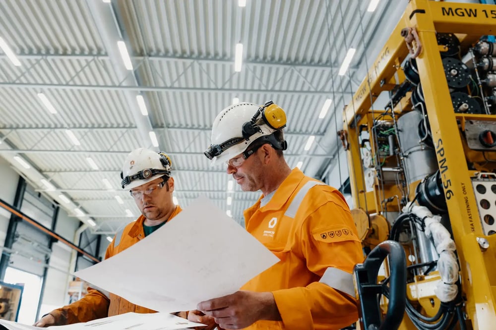 Two Enhanced Drilling engineers reviewing technical drawings beside subsea equipment in a workshop. Two Enhanced Drilling engineers reviewing technical drawings beside subsea equipment in a workshop.